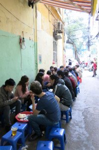 Small plastic chairs and tables lining the side of an alley, crowded with locals.